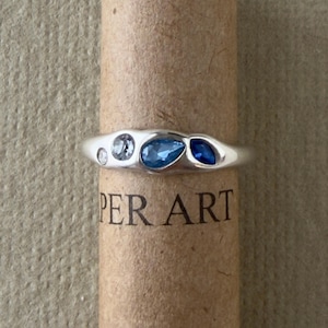 May include: A silver ring featuring four embedded stones in varying shades of blue and clear, arranged in a row. The stones include a small clear round, a larger light blue round, a medium blue teardrop, and a dark blue marquise shape.