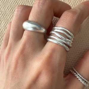 May include: A set of silver-toned rings worn on three fingers. One finger features a smooth, thick, rounded band. Another finger displays a stack of five thin, wavy, irregular bands. The third finger has two thin, twisted, rope-textured bands.