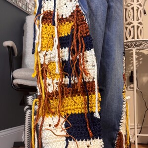 May include: A vibrant, hand-crocheted item with a patchwork design. It showcases horizontal stripes in white, navy, yellow, and brown, with dangling yarn and leather details. The piece is attached to a pair of blue jeans.