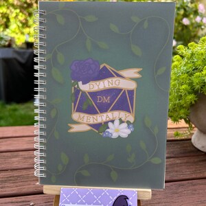 May include: A green spiral bound notebook featuring a purple and gold die with the words "Dying DM Mentally" on it. The die is surrounded by vines and flowers. The notebook is standing on a wooden easel with a business card that says "Phantom Min" and has a ghost with a witch hat on it.