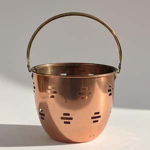 Pode incluir: Um balde de metal cor de cobre com uma alça de latão. O balde tem uma base arredondada e recortes retangulares nas laterais. A alça arqueia-se sobre a parte superior do balde, conectando-se em ambos os lados. Objeto decorativo.