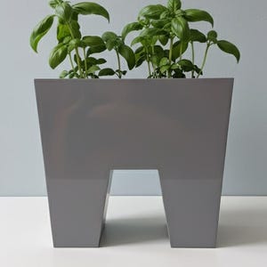 May include: A gray, modern planter with a unique cutout design, filled with vibrant green basil plants. The planter has a glossy finish and sits on a white surface, with a light blue background.