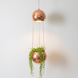 May include: A copper-colored, spherical hanging planter with a matching top sphere. The lower sphere holds green plants with trailing vines. The spheres are suspended by chains from a single point, creating a modern, minimalist design.
