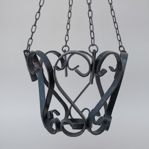 May include: A black metal hanging basket featuring curved, scroll-like ironwork patterns. The structure is suspended by three thin metal chains that meet at a single point at the top, creating a symmetrical, open-frame design for display.