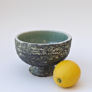 May include: A textured ceramic bowl with a dark green and black exterior and a lighter green interior. A yellow lemon sits next to the bowl. The bowl is on a pedestal base.