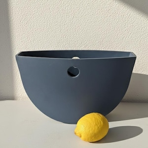 May include: A navy blue, semi-circular container with a circular opening. A bright yellow lemon sits next to the container. The container is on a white surface.
