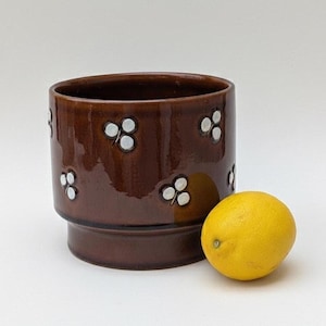 May include: A brown ceramic planter with white floral accents. The cylindrical pot has a wide opening and a raised base. A yellow lemon sits next to the planter. The planter is suitable for indoor or outdoor use.