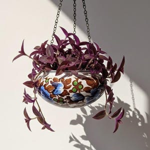 May include: A hanging planter with a trailing purple plant. The ceramic pot has a white base with a floral pattern in brown, blue, and green. The planter is suspended by a black chain against a white wall.