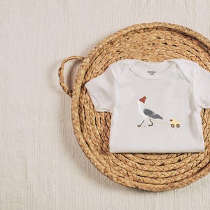 Könnte beinhalten: Weißer Baby-Strampler mit einem Aufdruck einer Möwe mit rotem Hut, die ein gelbes Spielzeugauto zieht.