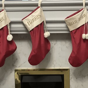 May include: Three red Christmas stockings with white pom-poms hanging on a white mantel above a fireplace. The stockings are personalised with the names "Phoebe", "Harris", and "Tillie-Anne" in gold lettering.