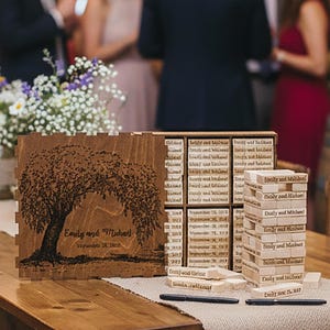 May include: A wooden table at a wedding reception with a decorative tree carving and a Jenga game. The tree carving has the names "Emily and Michael" and a date. Guests are in the background.