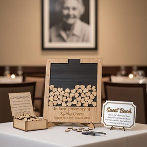 May include: A memorial guest book setup with a framed portrait, wooden sign, and heart-shaped tokens. The sign reads "In loving memory of Kathy Crone" with dates. A small box and a guest book sign are also present.