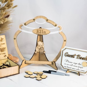 May include: Wooden guest book set with a Winnie the Pooh theme. The set includes a honey pot shaped stand with a clear acrylic insert, a small wooden box with a sign that says "Please sign your name so our sweet little hunny will know you came", and a white sign that says "Guest Book Please sign the so we will never forget our loved ones who came".