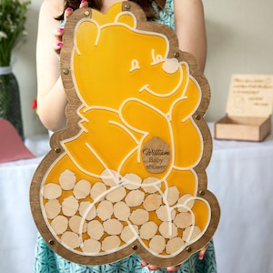 May include: A wooden Winnie the Pooh-themed baby shower guest book with a yellow Winnie the Pooh outline and a white inscription. The guest book has a wooden frame and a clear acrylic insert with a space for guests to write messages. The inscription reads "William Baby Shower".
