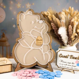 May include: A wooden Winnie the Pooh-shaped gender reveal sign with a clear acrylic insert. The sign is on a wooden stand, with pink and blue tokens, a small wooden box, and a guest book. The sign reads "Rhodes Gender Reveal."