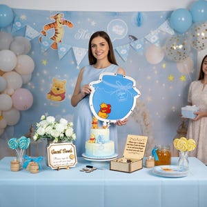 May include: A Winnie the Pooh themed party with a light blue color scheme. The scene includes balloons, a cake, and decorations. A woman holds a honey pot shaped sign with Winnie the Pooh characters. Other women hold gifts.