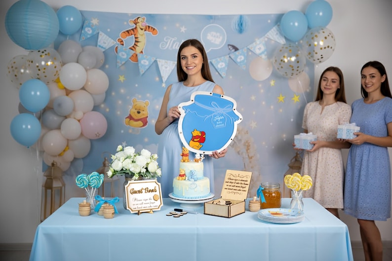 May include: A Winnie the Pooh themed party with a light blue color scheme. The scene includes balloons, a cake, and decorations. A woman holds a honey pot shaped sign with Winnie the Pooh characters. Other women hold gifts.