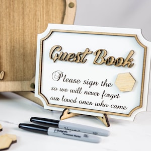May include: A wooden sign with the text "Guest Book" and the message "Please sign the so we will never forget our loved ones who came". The sign is on a stand and there are two black markers and wooden honeycomb shapes on a white surface.