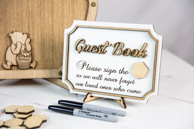 May include: A wooden sign with the text "Guest Book" and the message "Please sign the so we will never forget our loved ones who came". The sign is on a stand and there are two black markers and wooden honeycomb shapes on a white surface.