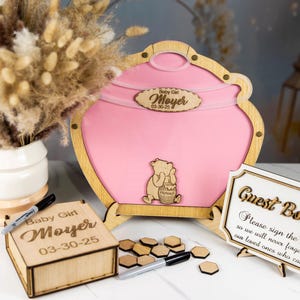 May include: Wooden guest book box with a Winnie the Pooh theme. The box is pink and has a Winnie the Pooh character on it. The box is decorated with a wooden sign that says "Guest Book" and a wooden sign that says "Please sign the so we will never forget our loved ones who came".