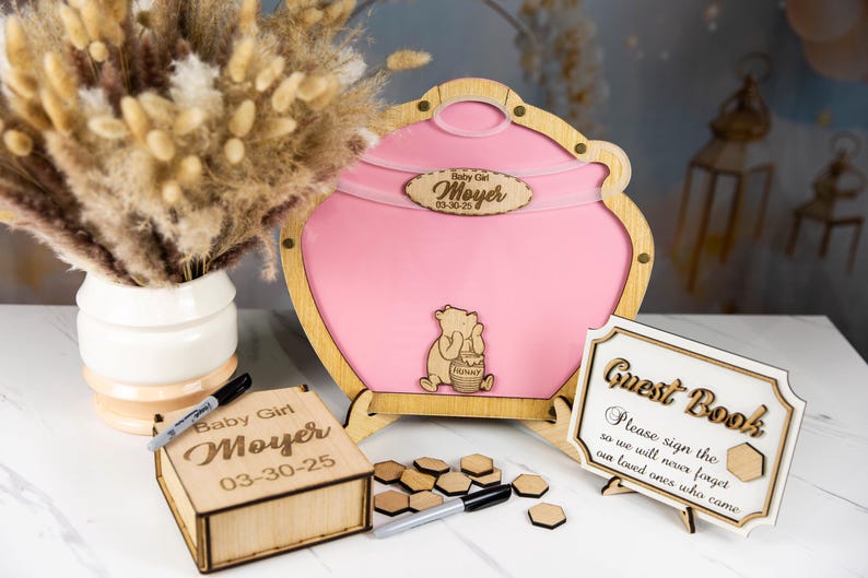 May include: Wooden guest book box with a Winnie the Pooh theme. The box is pink and has a Winnie the Pooh character on it. The box is decorated with a wooden sign that says "Guest Book" and a wooden sign that says "Please sign the so we will never forget our loved ones who came".