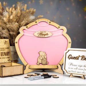 May include: A pink wooden honey pot guest book with a Winnie the Pooh character on the front. The honey pot is decorated with a wooden sign that says "Baby Girl Moyer 03.30.23". There is a small wooden box with a sign that says "Please sign your name so our sweet little hunny will know you came". There is also a sign that says "Guest Book Please sign the so we will never forget our loved ones who came".
