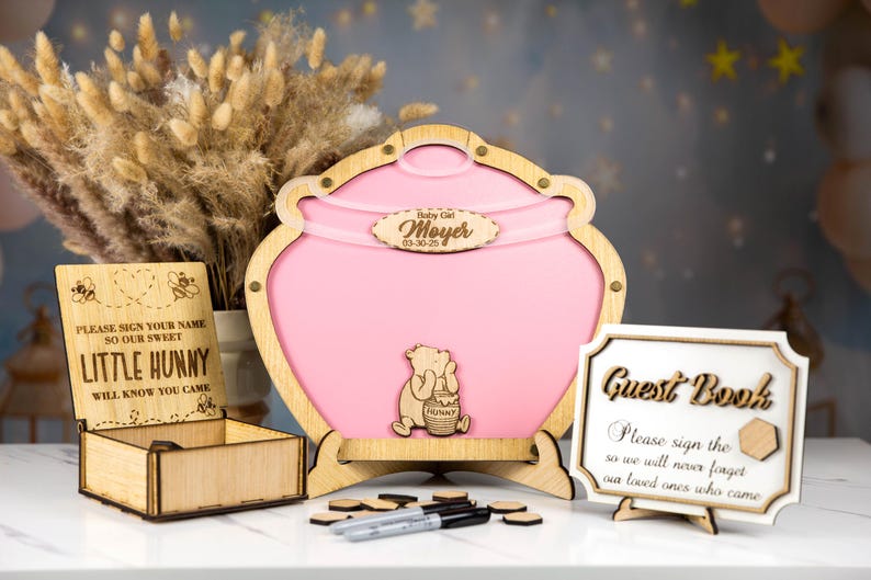 May include: A pink wooden honey pot guest book with a Winnie the Pooh character on the front. The honey pot is decorated with a wooden sign that says "Baby Girl Moyer 03.30.23". There is a small wooden box with a sign that says "Please sign your name so our sweet little hunny will know you came". There is also a sign that says "Guest Book Please sign the so we will never forget our loved ones who came".