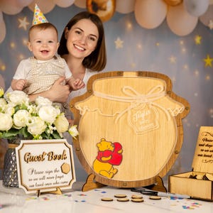 May include: A wooden Winnie the Pooh themed guest book set for a baby shower. The set includes a honey pot shaped guest book, a small box, and a sign that reads "Guest Book". A baby and a woman are in the background.