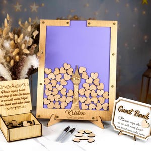 May include: A wooden guest book with a purple background and a silhouette of Rapunzel's tower. The guest book has a sign that says "Please sign a heart and drop it into the frame so we will never forget our loved ones who came." The sign also includes the name "Kristen" and the date "April 20, 2025".