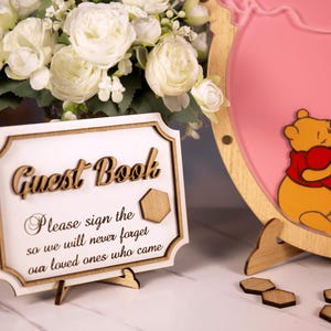 May include: A wooden guest book sign with the words "Guest Book" in a decorative font. The sign includes the text "Please sign the so we will never forget our loved ones who came." A pink decorative sign with a cartoon bear hugging a heart is also visible.