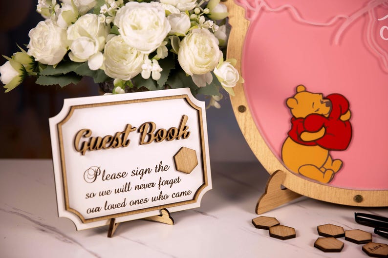 May include: A wooden guest book sign with the words "Guest Book" in a decorative font. The sign includes the text "Please sign the so we will never forget our loved ones who came." A pink decorative sign with a cartoon bear hugging a heart is also visible.