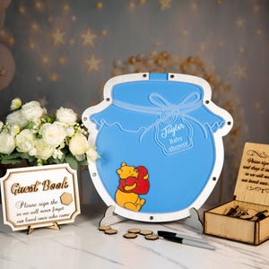May include: A baby shower guest book set featuring a blue honey pot-shaped sign with a Winnie the Pooh graphic, a wooden guest book sign, and a wooden box. The honey pot has the text "Taylor Baby shower".