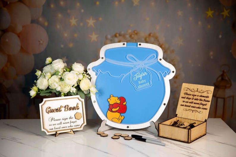 May include: A baby shower guest book set featuring a blue honey pot-shaped sign with a Winnie the Pooh graphic, a wooden guest book sign, and a wooden box. The honey pot has the text "Taylor Baby shower".