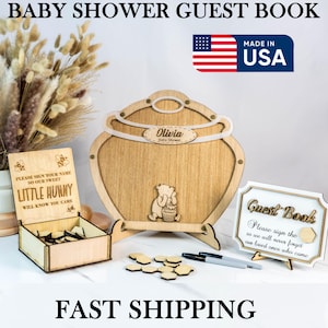 May include: Wooden baby shower guest book set. The set includes a large wooden guest book shaped like a honey pot, a smaller wooden box with guest sign-in pieces, and a sign. The guest book is engraved with the name "Olivia" and a Winnie the Pooh design.