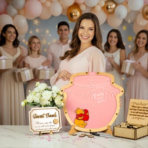 May include: A pink Winnie the Pooh themed guest book with the name "Emily" engraved on it. The guest book is surrounded by a group of people at a baby shower. A sign that says "Guest Book" is also on the table.
