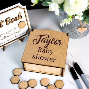May include: A wooden baby shower guest book box with the name "Taylor" engraved on the lid. A matching sign reads "Guest Book" and "Please sign the so we will never forget our loved ones who came."