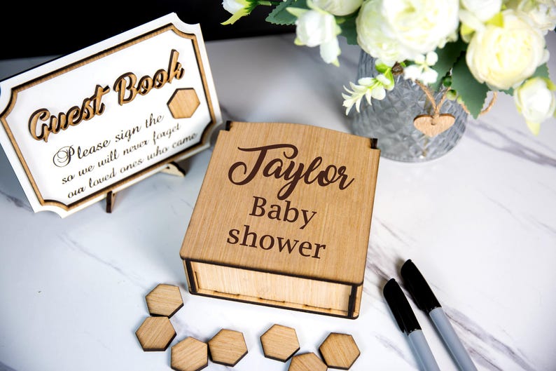 May include: A wooden baby shower guest book box with the name "Taylor" engraved on the lid. A matching sign reads "Guest Book" and "Please sign the so we will never forget our loved ones who came."