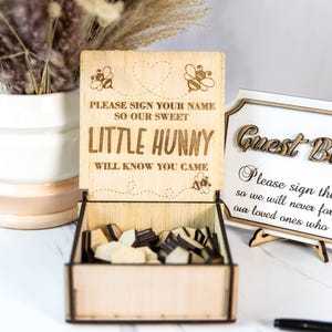 May include: Wooden guest book box with a sign that says "Please sign your name so our sweet little hunny will know you came." The box is open and filled with wooden hearts. A separate sign says "Guest Book" and "Please sign the so we will never forget our loved ones who came."