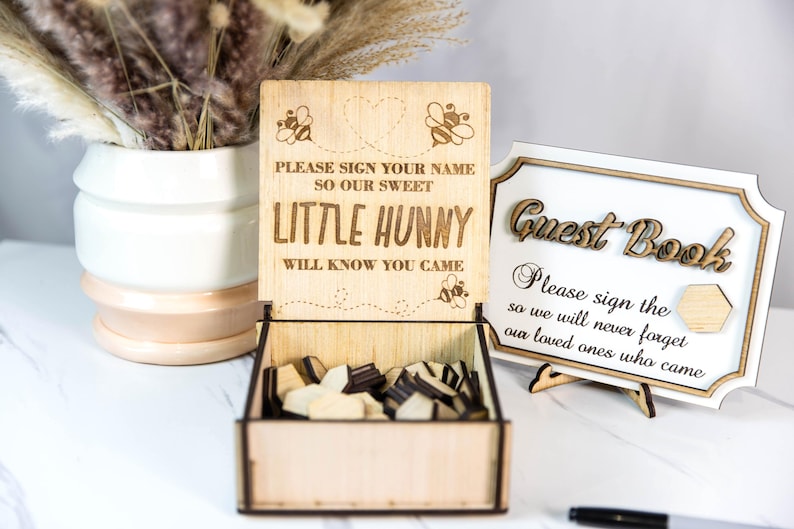 May include: Wooden guest book box with a sign that says "Please sign your name so our sweet little hunny will know you came." The box is open and filled with wooden hearts. A separate sign says "Guest Book" and "Please sign the so we will never forget our loved ones who came."