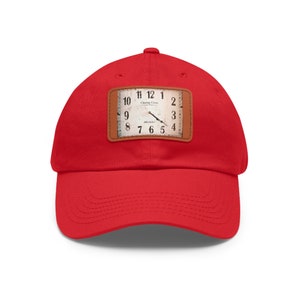 Könnte beinhalten: Eine leuchtend rote Baseballkappe mit einem rechteckigen Aufnäher mit einem Vintage-Zifferblatt. Das Zifferblatt hat schwarze Ziffern und Zeiger, mit dem Text "Charing Cross" sichtbar. Die Kappe ist aus weichem Material.