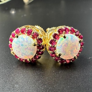 May include: A pair of gold earrings featuring a white opal centre stone surrounded by a halo of small red gemstones.