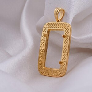 May include: A gold-colored pendant with a rectangular frame and a textured design. The pendant has a loop at the top for a chain and is displayed on a white, silky fabric. The pendant's dimensions are not visible.
