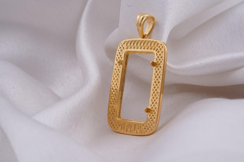 May include: A gold-colored pendant with a rectangular frame and a textured design. The pendant has a loop at the top for a chain and is displayed on a white, silky fabric. The pendant's dimensions are not visible.