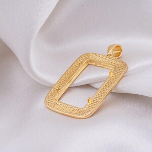 May include: A gold-colored rectangular pendant with a textured border and a loop at the top. The pendant is set against a white, silky background, creating a contrast that highlights its design and shape. The pendant's design is intricate.