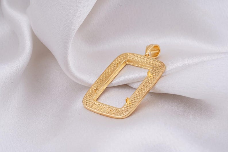 May include: A gold-colored rectangular pendant with a textured border and a loop at the top. The pendant is set against a white, silky background, creating a contrast that highlights its design and shape. The pendant's design is intricate.