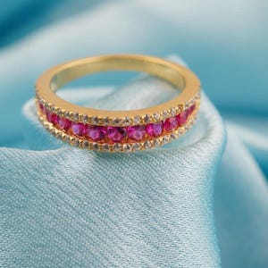 May include: A gold ring with a band of pink gemstones and a row of clear gemstones. The ring is set against a light blue fabric background.