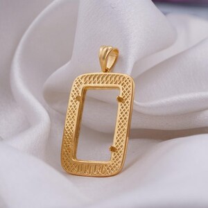 May include: A gold-colored rectangular pendant with an open center, resting on a white, silky surface. The pendant has a textured, patterned border and a small loop at the top for a chain. The overall design is simple yet elegant.