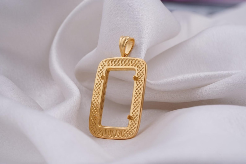 May include: A gold-colored rectangular pendant with an open center, resting on a white, silky surface. The pendant has a textured, patterned border and a small loop at the top for a chain. The overall design is simple yet elegant.