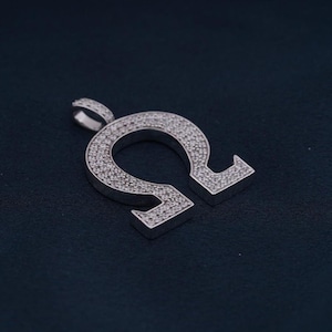 May include: A silver pendant in the shape of the Greek letter Omega, covered in small, sparkling stones. The pendant has a loop at the top for a chain and rests on a dark blue surface. The stones cover the entire surface of the Omega symbol.