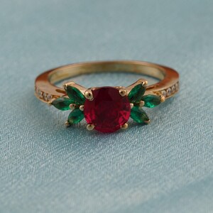 May include: A gold ring featuring a large, round, red gemstone as the focal point, flanked by green leaf-shaped stones. The band is also adorned with small, clear gemstones. The ring is set against a light blue background.